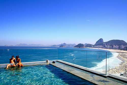 rio de janeiro A couple sitting on the edge of a swimming pool overlooking a crowded beach and a deep blue sea water